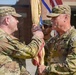Two U.S. Army soldiers in OCP (Operational Camouflage Pattern) uniforms are participating in a formal guidon transfer ceremony, likely a change of command or responsibility. The soldier on the right, identified by the name tape 'PARTIN' and wearing a First Cavalry Division patch, is passing or receiving a unit guidon to/from the soldier on the left. Additional soldiers in uniform are visible in the background holding flags.