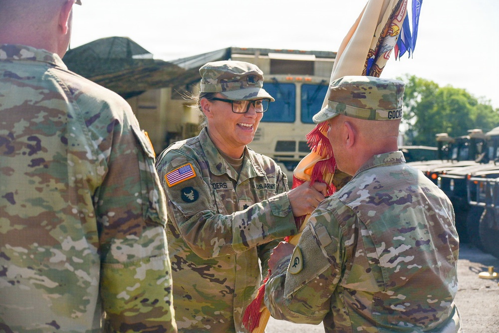 A U.S. Army ceremony is taking place outdoors, likely a change of command or unit activation event. A female soldier identified by nametape as 'DERS' (partially visible) is passing or receiving a unit guidon with red and gold fringe to/from a soldier identified as 'GOCKER'. Military vehicles and equipment are visible in the background.