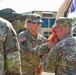 A U.S. Army ceremony is taking place outdoors, likely a change of command or unit activation event. A female soldier identified by nametape as 'DERS' (partially visible) is passing or receiving a unit guidon with red and gold fringe to/from a soldier identified as 'GOCKER'. Military vehicles and equipment are visible in the background.