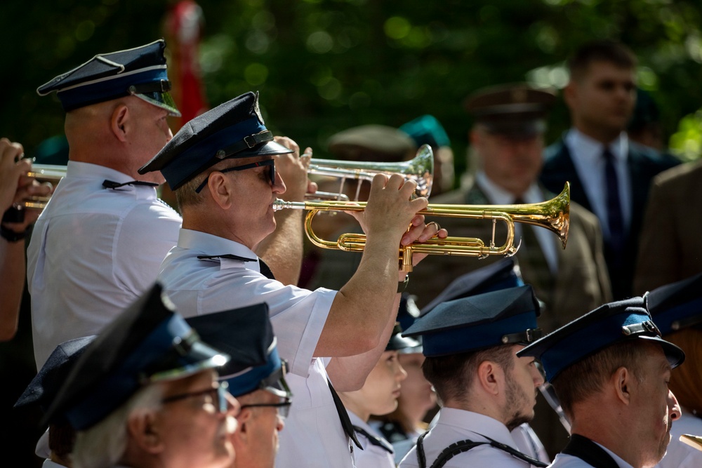 A military or ceremonial brass band is performing outdoors, with musicians dressed in white uniforms and dark blue peaked caps. The foreground features two prominent trumpet players actively playing their instruments. In the background, additional uniformed personnel and civilians in suits are visible, suggesting a formal public ceremony.