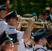 A military or ceremonial brass band is performing outdoors, with musicians dressed in white uniforms and dark blue peaked caps. The foreground features two prominent trumpet players actively playing their instruments. In the background, additional uniformed personnel and civilians in suits are visible, suggesting a formal public ceremony.