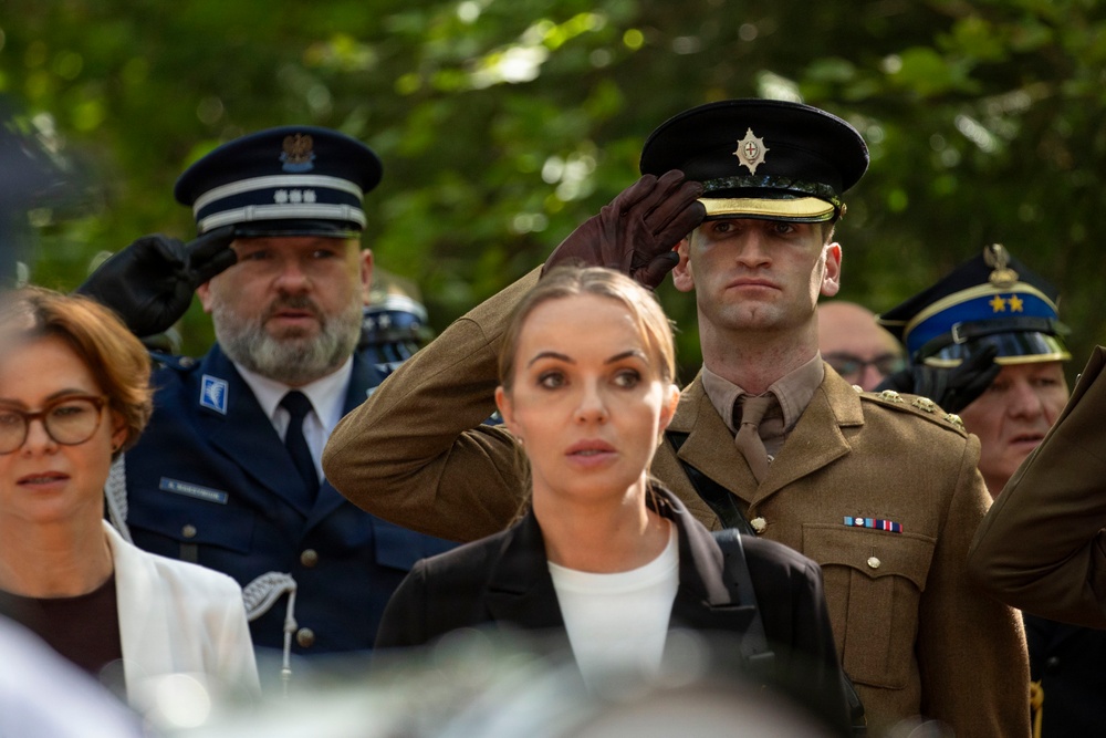 A formal military or ceremonial gathering showing multiple uniformed personnel and civilians. A soldier in an olive/brown military uniform with a peaked cap and service ribbons is performing a salute with a gloved hand. The scene appears to be an outdoor ceremony, possibly a memorial or official state event.