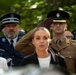 A formal military or ceremonial gathering showing multiple uniformed personnel and civilians. A soldier in an olive/brown military uniform with a peaked cap and service ribbons is performing a salute with a gloved hand. The scene appears to be an outdoor ceremony, possibly a memorial or official state event.