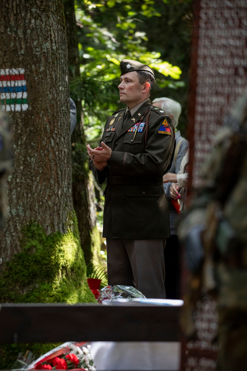 A U.S. Army officer in Army Service Uniform (ASU) stands at attention during what appears to be a memorial or commemorative ceremony in a forested outdoor setting. The officer wears the rank of Lieutenant Colonel (oak leaf cluster) and displays a 1st Armored Division patch with 'Old Ironsides' tab. Floral arrangements and what appears to be a memorial structure are visible in the foreground.