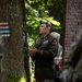 A U.S. Army officer in Army Service Uniform (ASU) stands at attention during what appears to be a memorial or commemorative ceremony in a forested outdoor setting. The officer wears the rank of Lieutenant Colonel (oak leaf cluster) and displays a 1st Armored Division patch with 'Old Ironsides' tab. Floral arrangements and what appears to be a memorial structure are visible in the foreground.