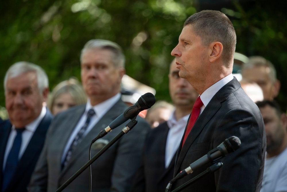 A man in a dark suit with a red tie stands at a microphone podium, appearing to deliver a speech at an outdoor public event. Several other formally dressed individuals stand in the background, suggesting an official or ceremonial gathering. The setting appears to be outdoors with green foliage visible in the background.
