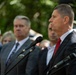 A man in a dark suit with a red tie stands at a microphone podium, appearing to deliver a speech at an outdoor public event. Several other formally dressed individuals stand in the background, suggesting an official or ceremonial gathering. The setting appears to be outdoors with green foliage visible in the background.