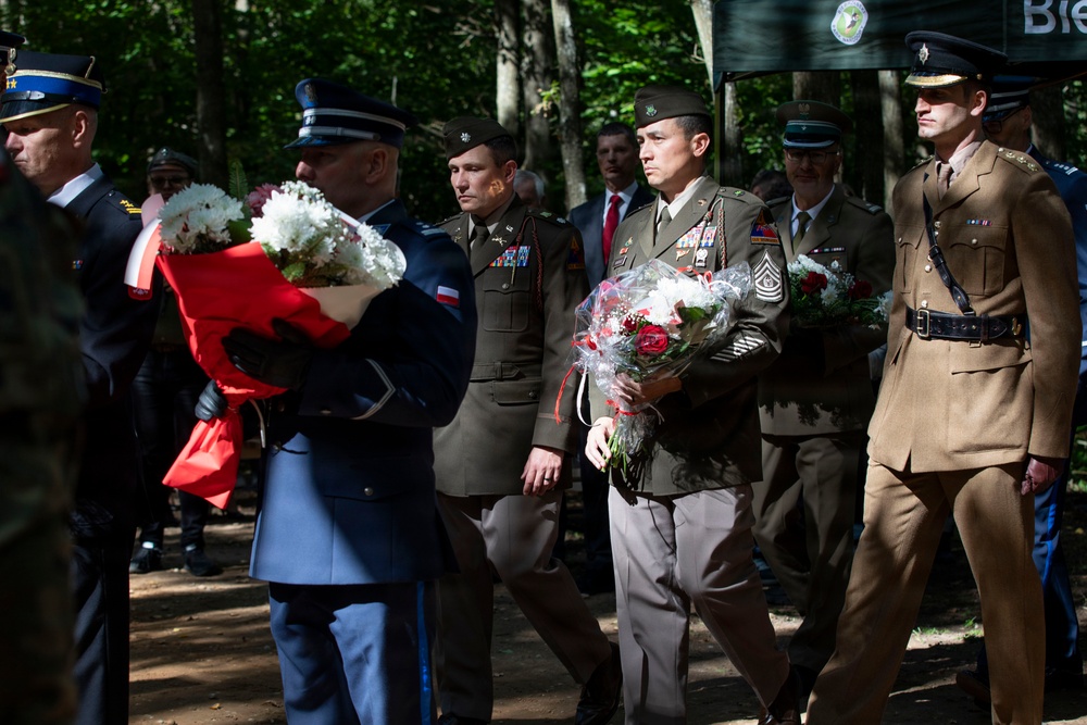 A formal military ceremony is taking place outdoors in a wooded setting, with personnel from multiple nations carrying floral wreaths and bouquets. The procession includes officers in distinct uniforms representing at least three different military branches or nations, including what appears to be US Army, US Air Force, and British Army personnel. The presence of red-and-white wrapped flowers suggests a possible Polish connection, indicating this may be a commemorative or memorial ceremony.