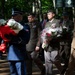 A formal military ceremony is taking place outdoors in a wooded setting, with personnel from multiple nations carrying floral wreaths and bouquets. The procession includes officers in distinct uniforms representing at least three different military branches or nations, including what appears to be US Army, US Air Force, and British Army personnel. The presence of red-and-white wrapped flowers suggests a possible Polish connection, indicating this may be a commemorative or memorial ceremony.