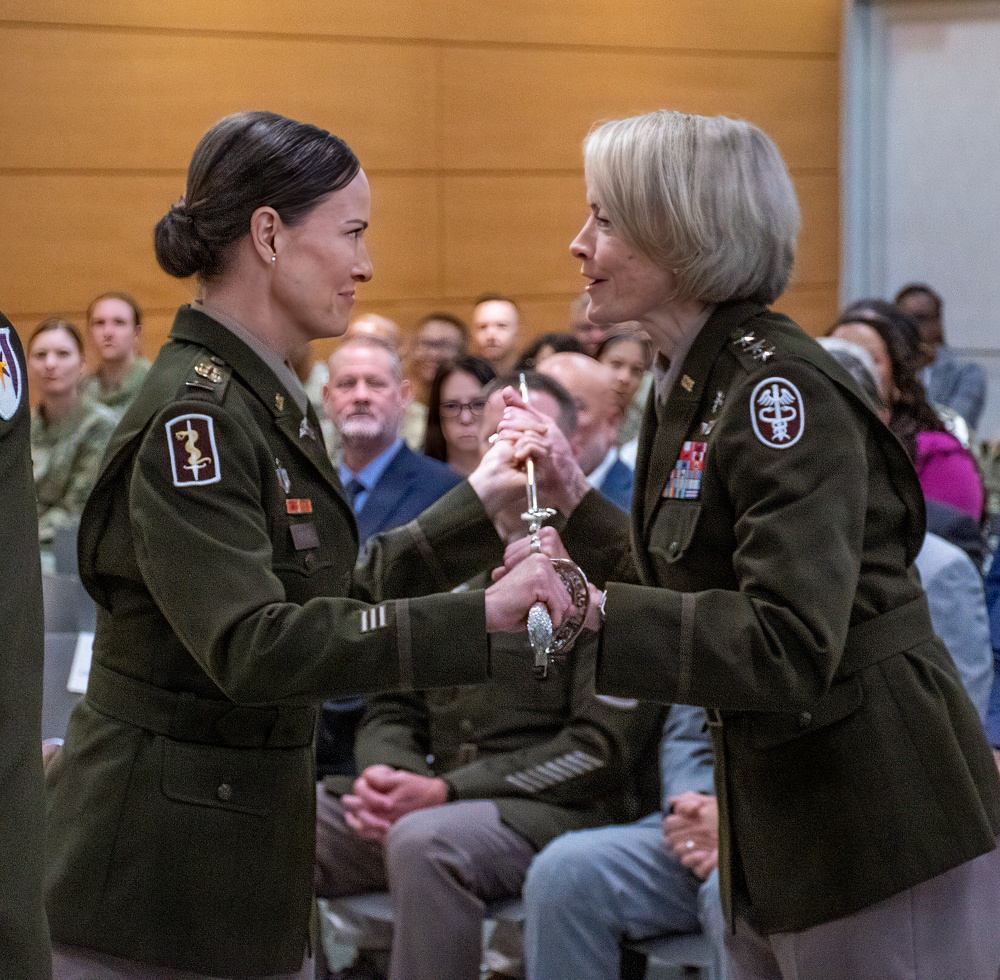 Two female U.S. Army Medical Corps officers are engaged in a formal military ceremony, appearing to exchange or present a ceremonial sword or saber. Both wear the Army Service Uniform (ASU) in olive green with Medical Corps insignia patches on their shoulders. An audience of military and civilian personnel is seated in the background observing the ceremony.