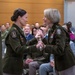 Two female U.S. Army Medical Corps officers are engaged in a formal military ceremony, appearing to exchange or present a ceremonial sword or saber. Both wear the Army Service Uniform (ASU) in olive green with Medical Corps insignia patches on their shoulders. An audience of military and civilian personnel is seated in the background observing the ceremony.