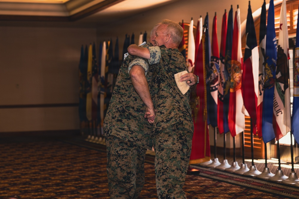 Two individuals wearing U.S. Marine Corps MARPAT digital camouflage uniforms are embracing in what appears to be a formal military ceremony or event. They are standing in front of a row of state and organizational flags displayed on stands. The setting appears to be an indoor banquet or conference hall.
