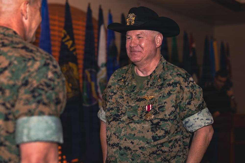 A U.S. Navy servicemember wearing digital camouflage utilities is shown at what appears to be a military ceremony or award presentation. The individual is wearing a distinctive black cowboy hat with a gold emblem and has a medal pinned to their uniform. Multiple military flags are visible in the background, suggesting a formal indoor military setting.