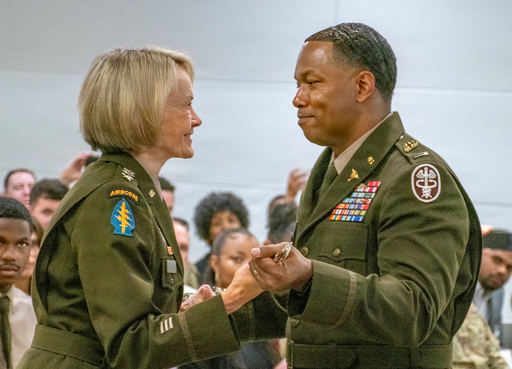 Two U.S. Army officers in Army Green Service Uniforms are engaged in what appears to be a military ceremony, likely a promotion or award presentation. The female officer on the left is pinning something onto or presenting something to the male officer on the right, who displays numerous service ribbons and a Medical Corps insignia. An audience of military and civilian personnel is visible in the background.