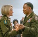 Two U.S. Army officers in Army Green Service Uniforms are engaged in what appears to be a military ceremony, likely a promotion or award presentation. The female officer on the left is pinning something onto or presenting something to the male officer on the right, who displays numerous service ribbons and a Medical Corps insignia. An audience of military and civilian personnel is visible in the background.