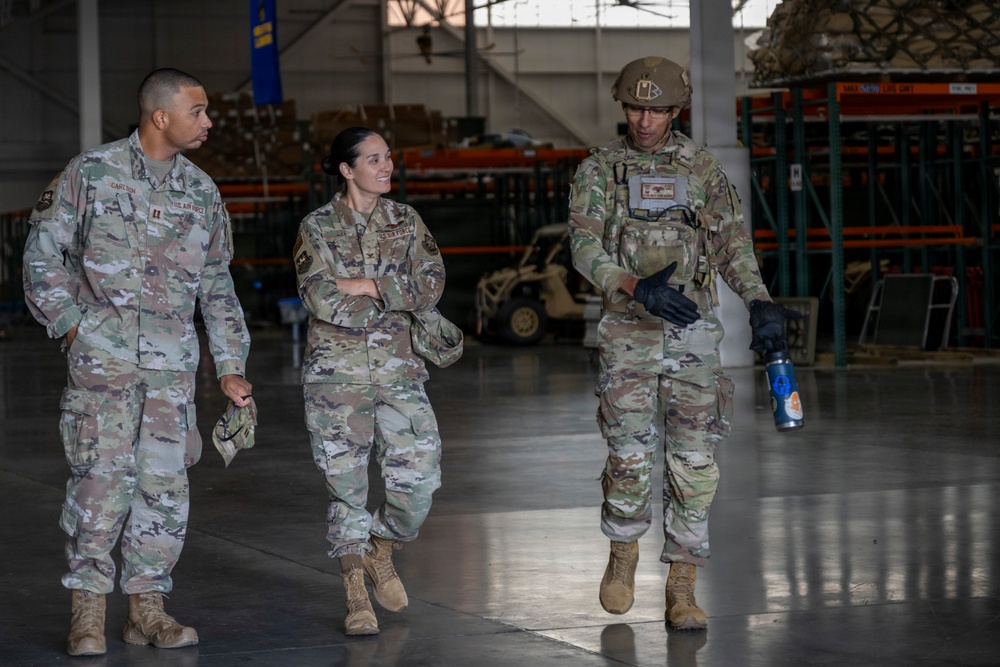 Three U.S. Air Force personnel in OCP (Operational Camouflage Pattern) uniforms are walking and conversing inside what appears to be a military warehouse or hangar facility. The individual on the right is wearing a helmet and tactical vest, suggesting a more operationally equipped role, while the other two are in standard duty uniforms. The setting includes visible storage shelving and logistics equipment in the background.