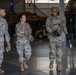 Three U.S. Air Force personnel in OCP (Operational Camouflage Pattern) uniforms are walking and conversing inside what appears to be a military warehouse or hangar facility. The individual on the right is wearing a helmet and tactical vest, suggesting a more operationally equipped role, while the other two are in standard duty uniforms. The setting includes visible storage shelving and logistics equipment in the background.