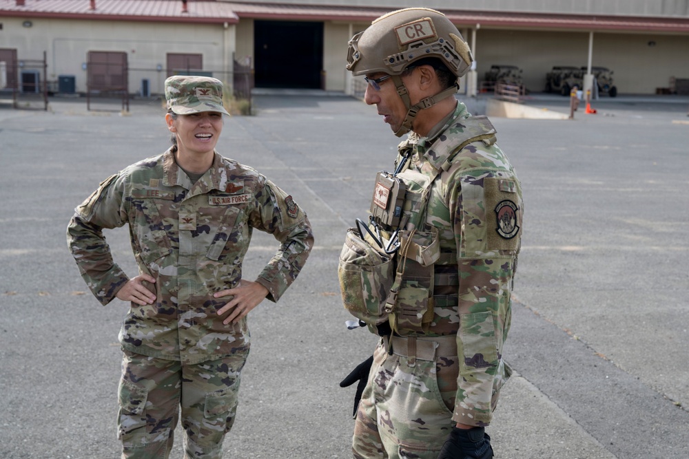 Two U.S. Air Force personnel are conversing on a military installation tarmac or parade ground. The individual on the left wears a standard OCP uniform with a colonel's eagle rank insignia and a 'U.S. AIR FORCE' tape, while the individual on the right is in full tactical gear including a ballistic helmet marked 'CR', body armor, and tactical vest. A large hangar or maintenance building is visible in the background along with military vehicles.