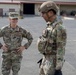 Two U.S. Air Force personnel are conversing on a military installation tarmac or parade ground. The individual on the left wears a standard OCP uniform with a colonel's eagle rank insignia and a 'U.S. AIR FORCE' tape, while the individual on the right is in full tactical gear including a ballistic helmet marked 'CR', body armor, and tactical vest. A large hangar or maintenance building is visible in the background along with military vehicles.