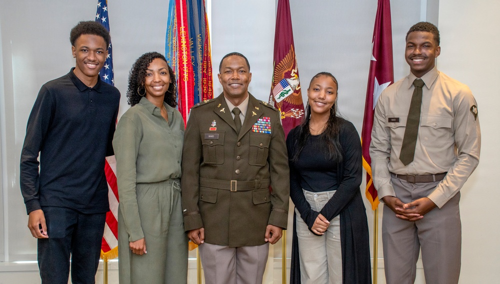 A formal group portrait of five individuals posing in front of multiple military and American flags. The central figure is wearing a U.S. Army dress uniform with multiple service ribbons and decorations, flanked by four civilians. The setting appears to be an official military office or ceremony room.