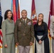 A formal group portrait of five individuals posing in front of multiple military and American flags. The central figure is wearing a U.S. Army dress uniform with multiple service ribbons and decorations, flanked by four civilians. The setting appears to be an official military office or ceremony room.