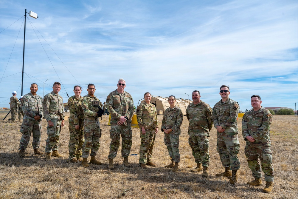 A group of ten military personnel in U.S. Army Operational Camouflage Pattern (OCP) uniforms pose together for a group photo outdoors on a dry grass field. Behind them are tan military field tents and a communications pole with guy-wires, suggesting a field exercise or training environment. The setting appears to be a military installation or training area.