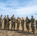 A group of ten military personnel in U.S. Army Operational Camouflage Pattern (OCP) uniforms pose together for a group photo outdoors on a dry grass field. Behind them are tan military field tents and a communications pole with guy-wires, suggesting a field exercise or training environment. The setting appears to be a military installation or training area.