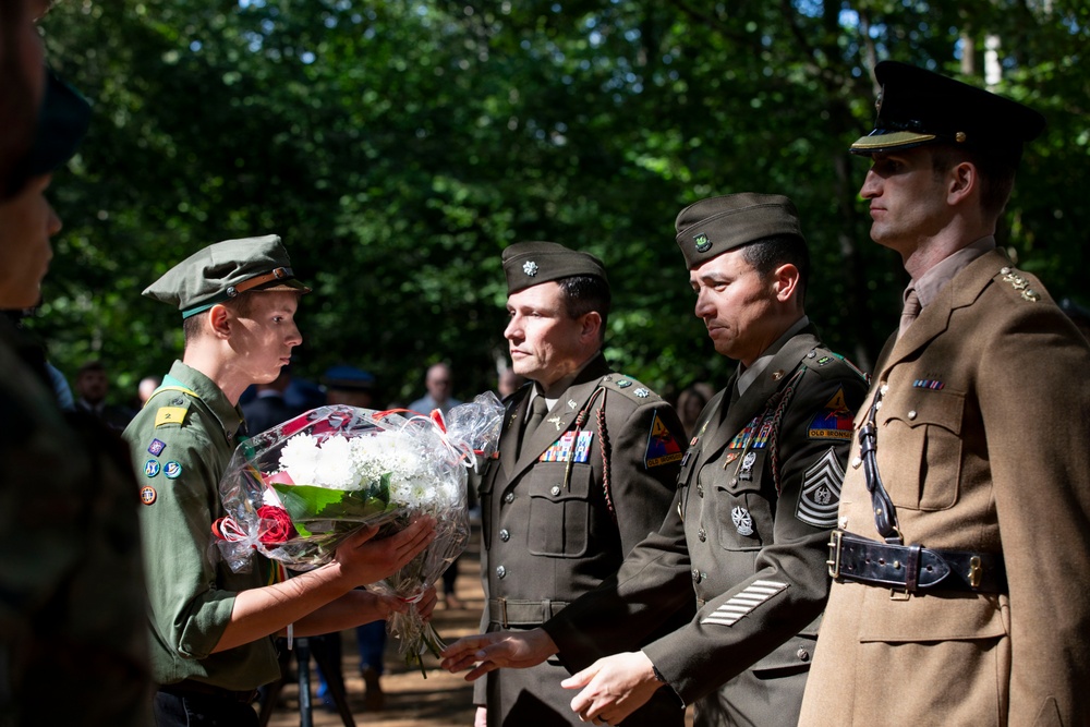 A ceremonial event appears to be taking place outdoors in a wooded setting, where a young person in a scout-style uniform is presenting or receiving a bouquet of flowers to/from military personnel in olive drab uniforms. The military figures are wearing decorated dress uniforms with medals, rank insignia, and garrison caps. The scene suggests a commemorative or memorial ceremony.