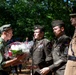 A ceremonial event appears to be taking place outdoors in a wooded setting, where a young person in a scout-style uniform is presenting or receiving a bouquet of flowers to/from military personnel in olive drab uniforms. The military figures are wearing decorated dress uniforms with medals, rank insignia, and garrison caps. The scene suggests a commemorative or memorial ceremony.