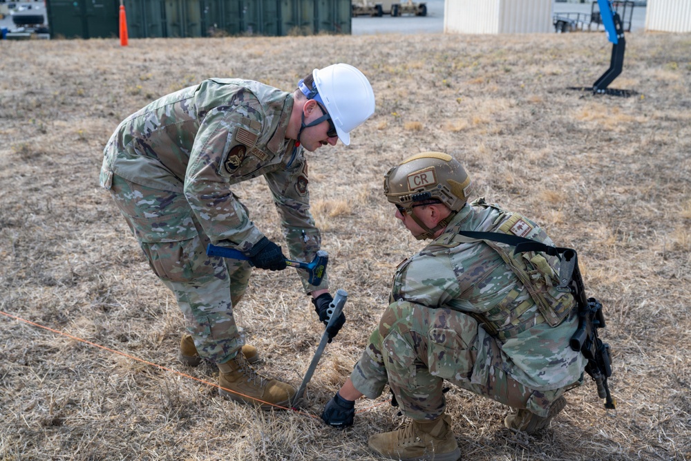 Two military personnel in OCP (Operational Camouflage Pattern) uniforms are working together on a dry grass field, with one driving a stake or anchor into the ground using a hammer while the other kneels to assist or observe. The standing soldier wears a white hard hat and safety glasses, while the kneeling soldier wears a tan tactical helmet marked 'CR'. They appear to be conducting a field setup or construction task at a military installation.