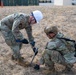 Two military personnel in OCP (Operational Camouflage Pattern) uniforms are working together on a dry grass field, with one driving a stake or anchor into the ground using a hammer while the other kneels to assist or observe. The standing soldier wears a white hard hat and safety glasses, while the kneeling soldier wears a tan tactical helmet marked 'CR'. They appear to be conducting a field setup or construction task at a military installation.