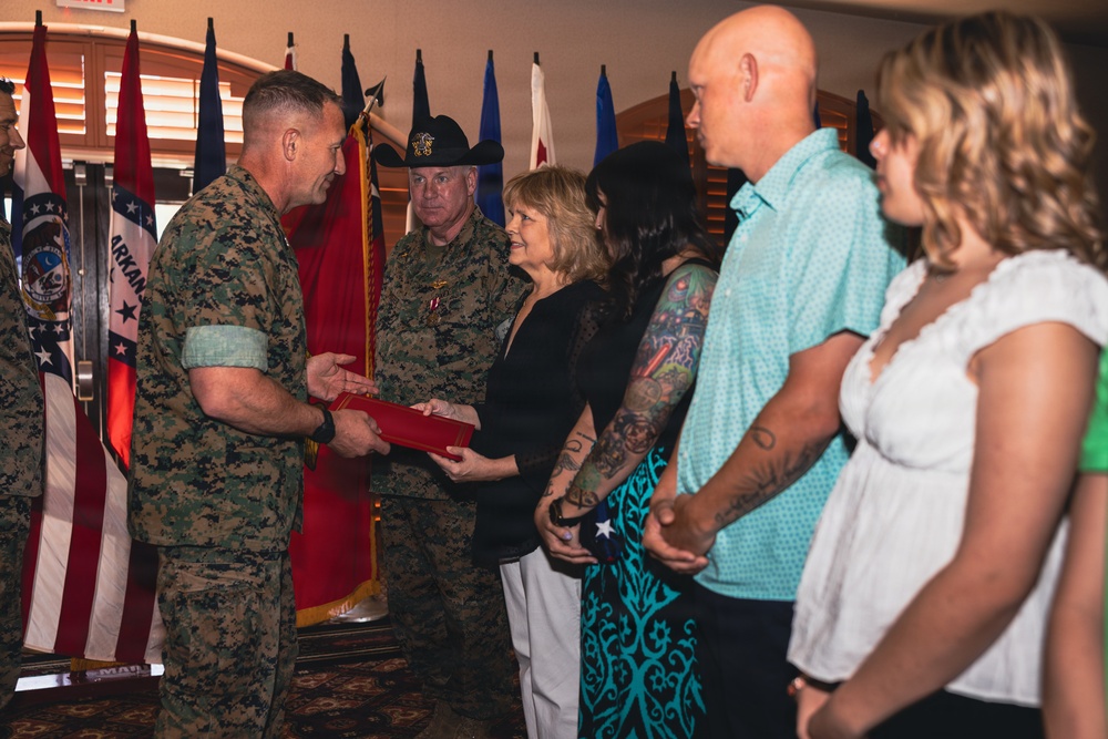 A U.S. Marine Corps officer in digital camouflage uniform is presenting a red folder or award case to a civilian woman during what appears to be a formal military ceremony. A second military figure wearing a cowboy hat and dress whites with medals stands nearby, alongside a group of civilians. Multiple military unit flags and state flags are displayed in the background.