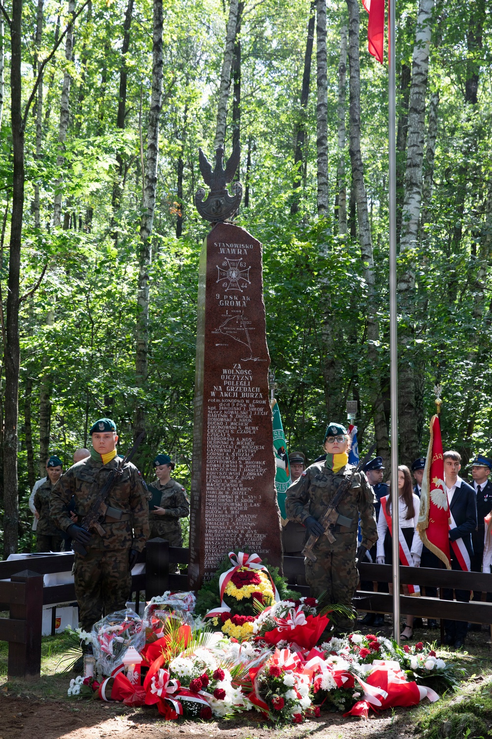 A military memorial ceremony is taking place in a forested area around a tall dark red granite obelisk monument. The monument bears the inscription 'STANOWISKO WAWRA' and references dates 1863 and 1944, along with '9 PSK AK GROMA' and 'ZA WOLNOŚĆ OJCZYZNY POLEGLI NA GRZĘDACH W AKCJI BURZA' (For the Freedom of the Fatherland, Fallen at Grzędy in Operation Tempest). Soldiers in Polish military uniforms stand guard while civilians and honor guard members with flags attend the commemoration.
