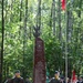A military memorial ceremony is taking place in a forested area around a tall dark red granite obelisk monument. The monument bears the inscription 'STANOWISKO WAWRA' and references dates 1863 and 1944, along with '9 PSK AK GROMA' and 'ZA WOLNOŚĆ OJCZYZNY POLEGLI NA GRZĘDACH W AKCJI BURZA' (For the Freedom of the Fatherland, Fallen at Grzędy in Operation Tempest). Soldiers in Polish military uniforms stand guard while civilians and honor guard members with flags attend the commemoration.