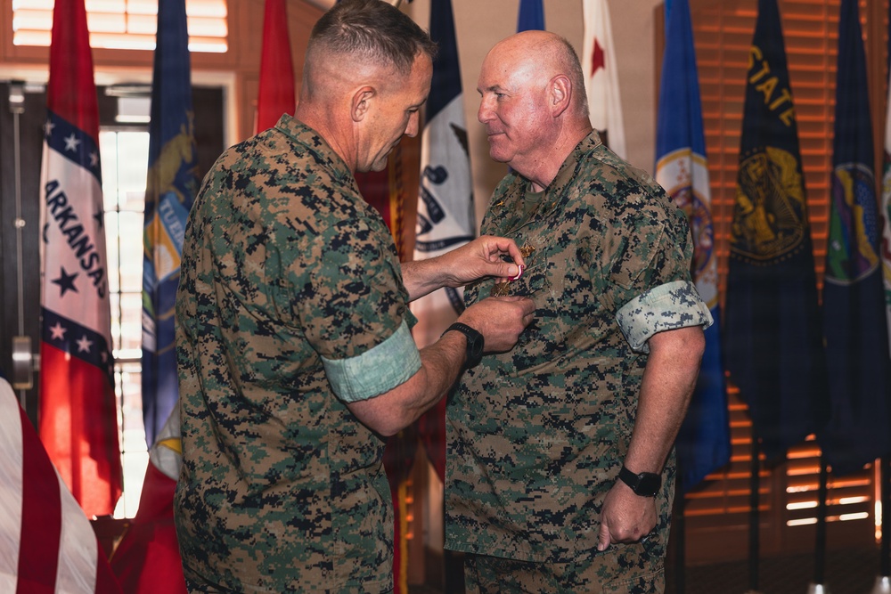 Two U.S. Marine Corps personnel in MARPAT digital camouflage uniforms are participating in a formal military awards ceremony. One Marine is pinning a medal onto the chest of the other Marine, who is standing at attention. The ceremony is taking place indoors in front of a display of multiple U.S. state and military flags.
