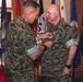 Two U.S. Marine Corps personnel in MARPAT digital camouflage uniforms are participating in a formal military awards ceremony. One Marine is pinning a medal onto the chest of the other Marine, who is standing at attention. The ceremony is taking place indoors in front of a display of multiple U.S. state and military flags.
