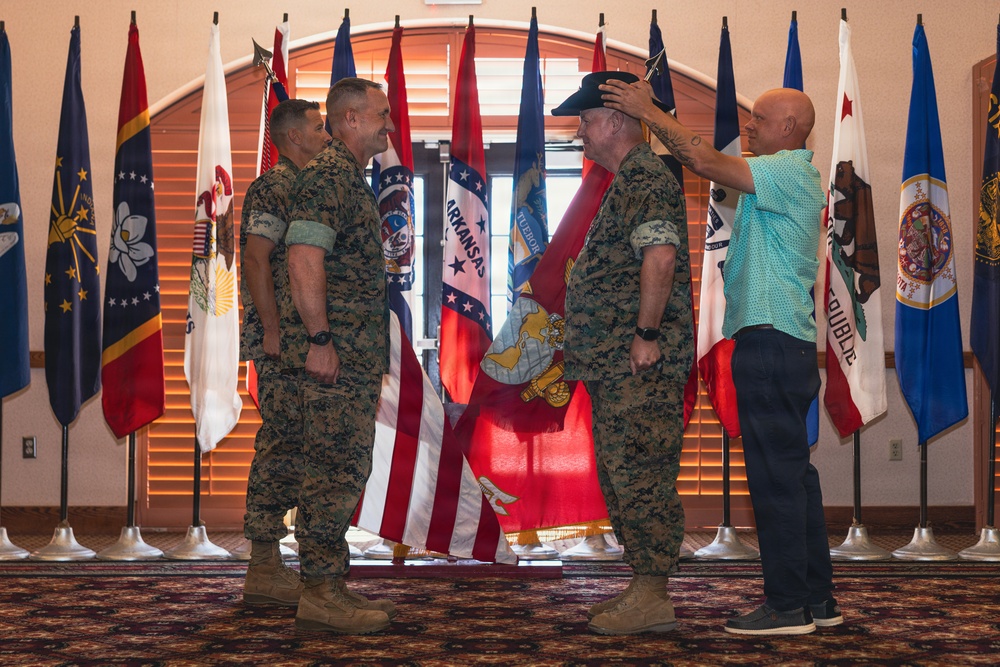A military ceremony is taking place in a formal hall lined with U.S. state flags. Three individuals in U.S. Marine Corps MARPAT camouflage uniforms are present alongside a civilian in a teal shirt, who appears to be placing a black cap on one of the Marines. The setting and interaction suggest a retirement or promotion ceremony.