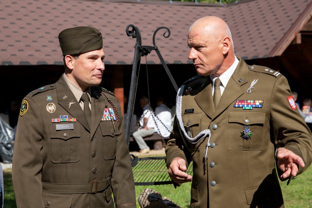 Two military officers in olive/khaki dress uniforms are engaged in conversation outdoors near a building. The officer on the left wears a U.S. Army uniform with a garrison cap and name tape reading 'SAXON', while the officer on the right wears what appears to be a Polish Army uniform with two-star rank insignia and a name tape reading 'WOJCK'. Both display multiple service ribbons and decorations.