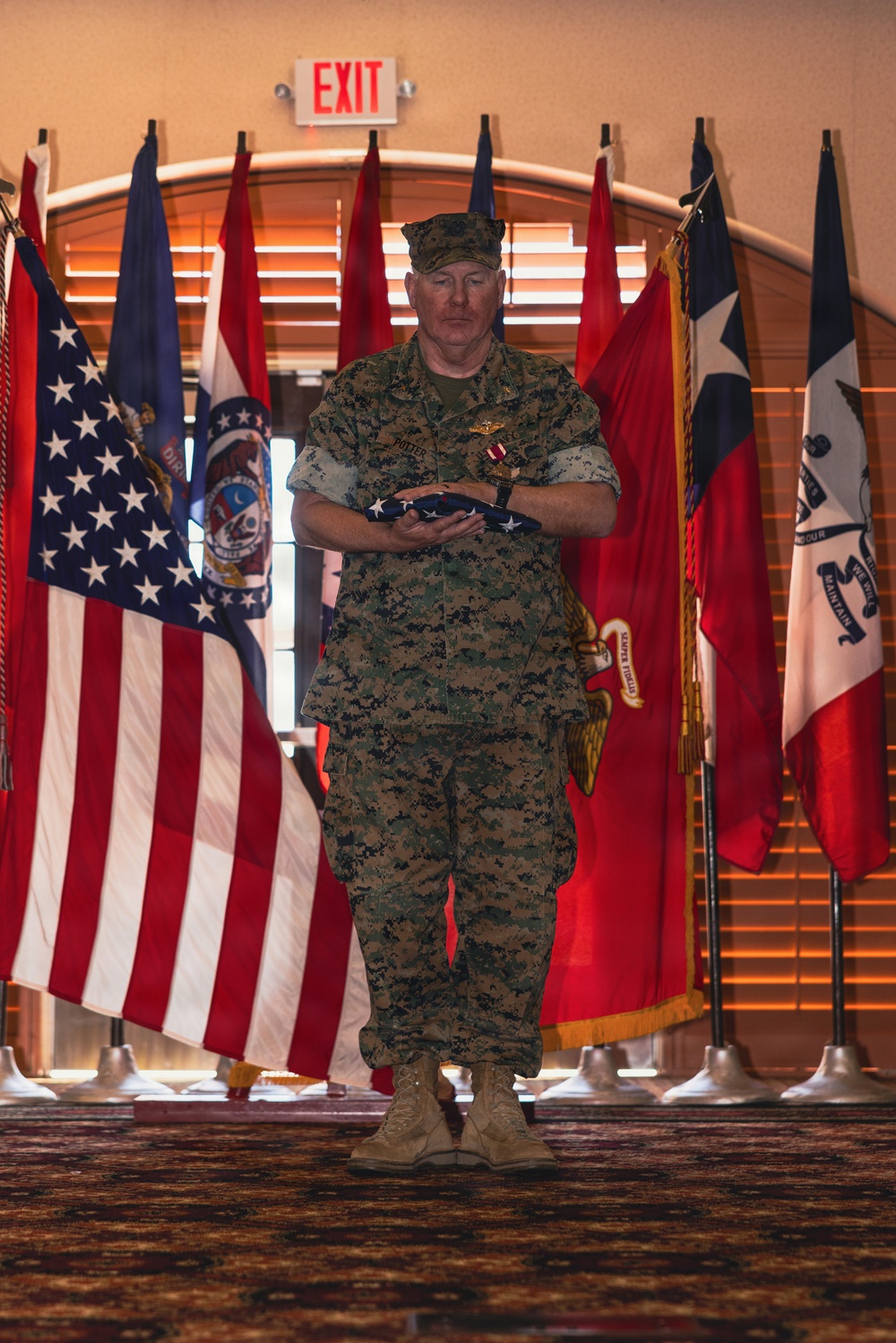 A U.S. Marine Corps service member in MARPAT digital camouflage uniform stands at attention holding a folded American flag during what appears to be a military retirement or awards ceremony. The individual, whose name tape reads 'POTTER,' is wearing a medal on their chest and a military aviator badge, standing before a display of multiple flags in a formal indoor venue. The setting suggests an official military ceremony with state and unit flags arranged ceremonially in the background.
