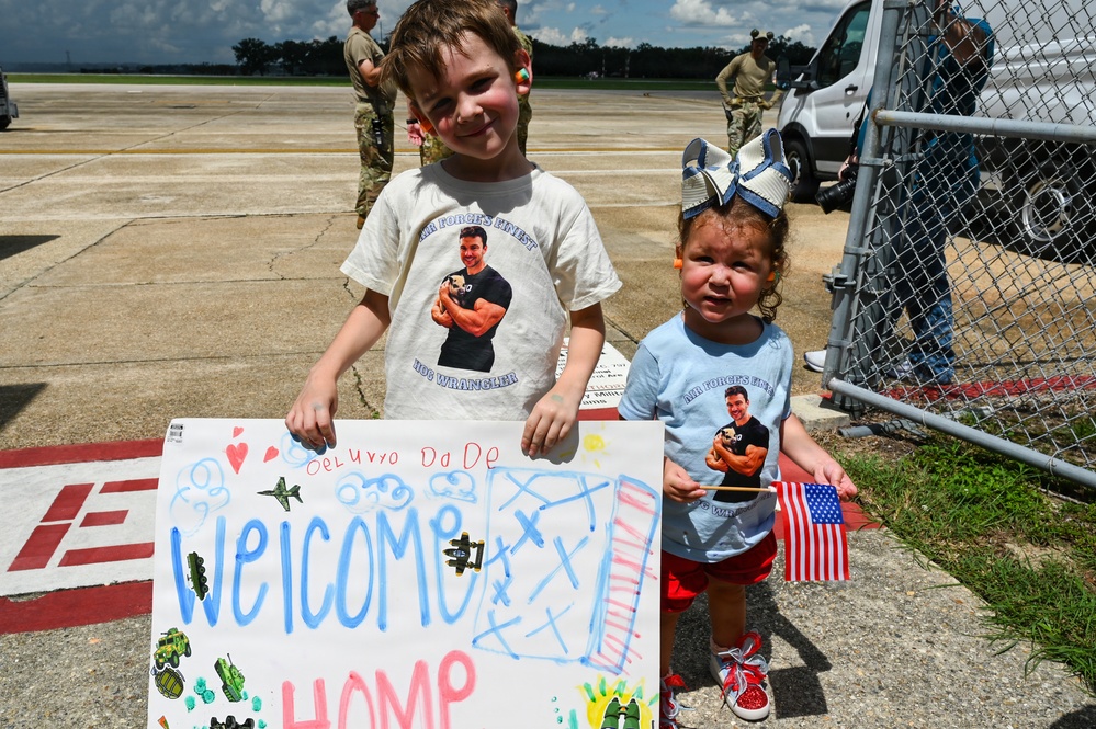Two young children stand on a military airfield tarmac holding a handmade 'Welcome Home' sign and an American flag, awaiting the return of a family member. Both children are wearing matching custom t-shirts featuring a photo of a muscular man labeled 'Air Force's Finest Hog Wrangler.' The scene depicts a military homecoming event with personnel and vehicles visible in the background.