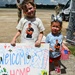 Two young children stand on a military airfield tarmac holding a handmade 'Welcome Home' sign and an American flag, awaiting the return of a family member. Both children are wearing matching custom t-shirts featuring a photo of a muscular man labeled 'Air Force's Finest Hog Wrangler.' The scene depicts a military homecoming event with personnel and vehicles visible in the background.