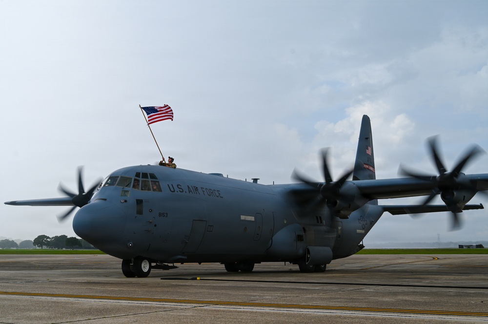 A U.S. Air Force C-130 Hercules military transport aircraft is taxiing on a flight line apron. A person is visible on top of the fuselage waving a large American flag. The aircraft's four turboprop engines are running, as evidenced by the motion blur on the propellers.