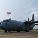A U.S. Air Force C-130 Hercules military transport aircraft is taxiing on a flight line apron. A person is visible on top of the fuselage waving a large American flag. The aircraft's four turboprop engines are running, as evidenced by the motion blur on the propellers.