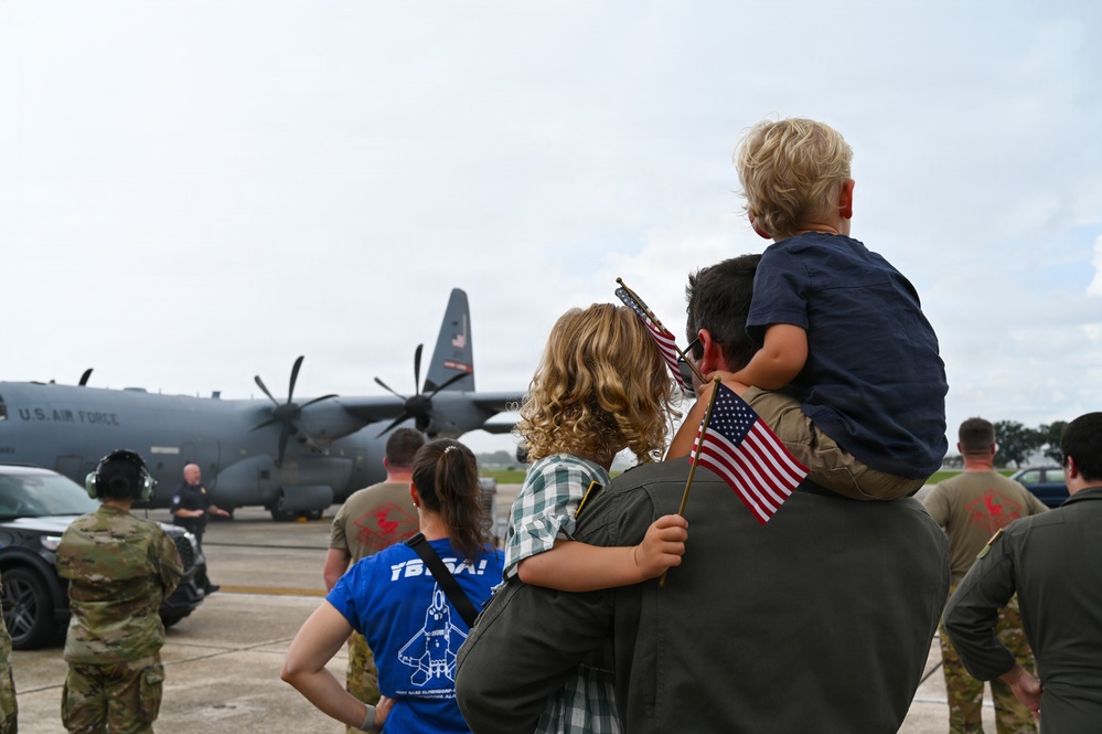 A military homecoming or sendoff ceremony on an airfield tarmac, with family members holding small American flags while watching a U.S. Air Force C-130 Hercules turboprop aircraft. A central figure carries a young child on their shoulders while embracing another person, surrounded by military personnel in flight suits and camouflage uniforms. The scene conveys an emotional military family reunion or departure event.
