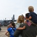 A military homecoming or sendoff ceremony on an airfield tarmac, with family members holding small American flags while watching a U.S. Air Force C-130 Hercules turboprop aircraft. A central figure carries a young child on their shoulders while embracing another person, surrounded by military personnel in flight suits and camouflage uniforms. The scene conveys an emotional military family reunion or departure event.