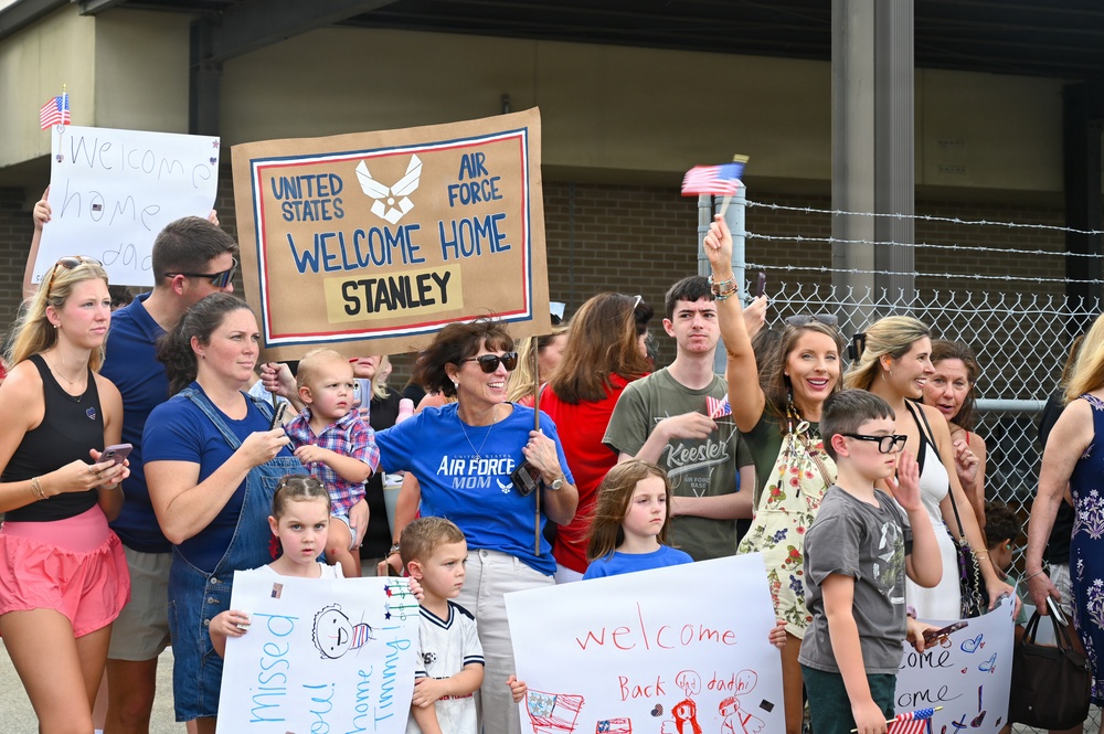 A crowd of families and loved ones are gathered at what appears to be a military base to welcome home United States Air Force service members. Multiple handmade signs are visible, including one reading 'United States Air Force Welcome Home Stanley' and others saying 'Welcome Back Daddy' and 'Welcome Home Timmy.' The group includes adults, teenagers, and young children, many waving American flags and holding signs in a celebratory homecoming event.