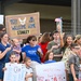 A crowd of families and loved ones are gathered at what appears to be a military base to welcome home United States Air Force service members. Multiple handmade signs are visible, including one reading 'United States Air Force Welcome Home Stanley' and others saying 'Welcome Back Daddy' and 'Welcome Home Timmy.' The group includes adults, teenagers, and young children, many waving American flags and holding signs in a celebratory homecoming event.