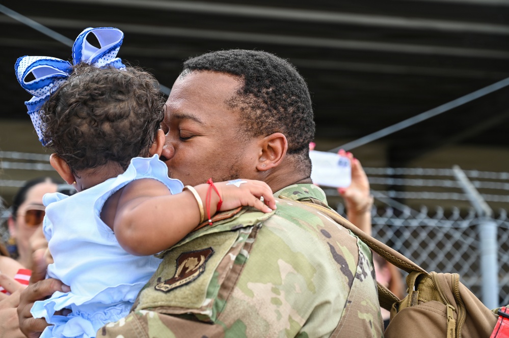 A military service member in OCP (Operational Camouflage Pattern) uniform is kissing a young infant who is dressed in a light blue outfit with a blue bow in her hair. The moment appears to be an emotional reunion or farewell at what looks like a military installation, with a chain-link fence and other people visible in the background. The image captures an intimate family moment between the soldier and the baby.