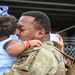 A military service member in OCP (Operational Camouflage Pattern) uniform is kissing a young infant who is dressed in a light blue outfit with a blue bow in her hair. The moment appears to be an emotional reunion or farewell at what looks like a military installation, with a chain-link fence and other people visible in the background. The image captures an intimate family moment between the soldier and the baby.