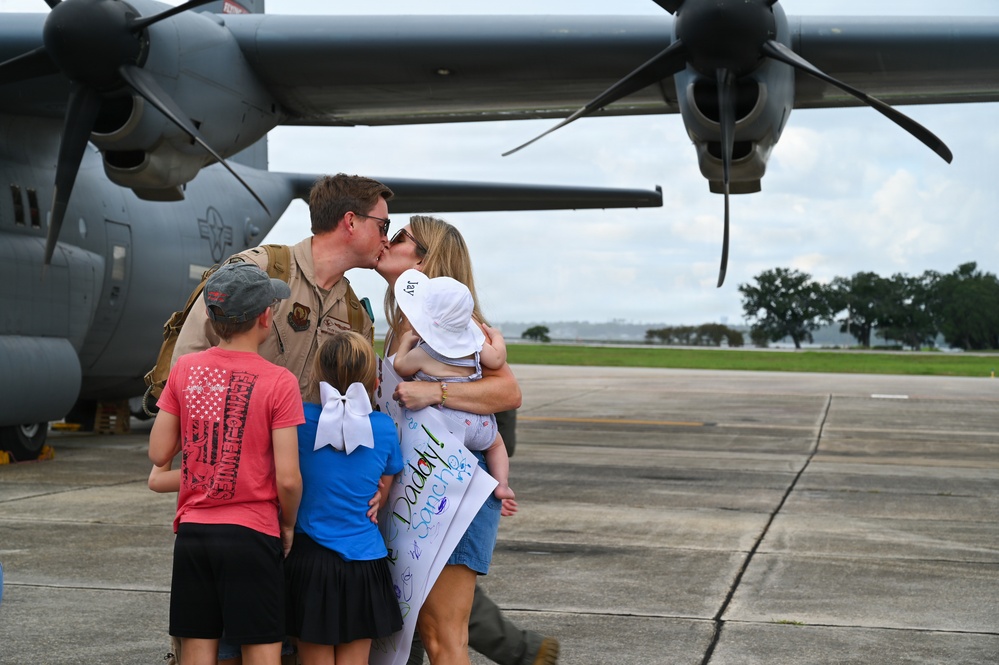 A military servicemember in flight suit is reuniting with his family on an airfield tarmac, sharing a kiss with a woman who is holding a baby. Two children stand nearby, and a handmade welcome home sign reading 'Daddy' is visible. A large military turboprop aircraft, consistent with a C-130 variant, is parked directly behind them.